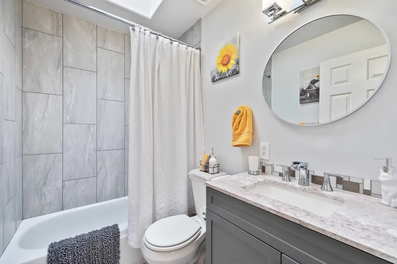 A modern bathroom with a skylight, gray tiled shower, a round mirror, and a white vanity in a Caldwell, Idaho home.