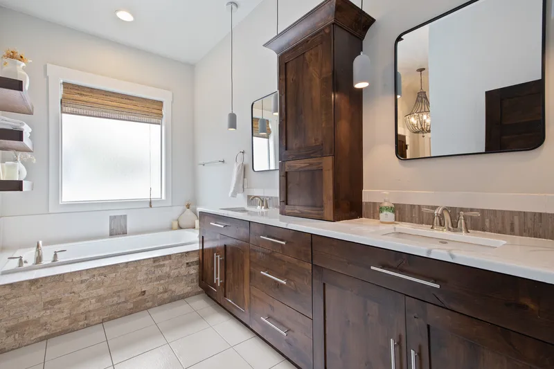 A luxury bathroom in Caldwell with dark wood cabinetry, a white double vanity, a built-in bathtub with stone accents, and modern lighting fixtures, captured for high-quality real estate photography.