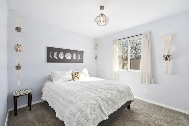 A modern boho-style bedroom with a white bed, macramé wall decor, a large window, and moon phase artwork above the bed.