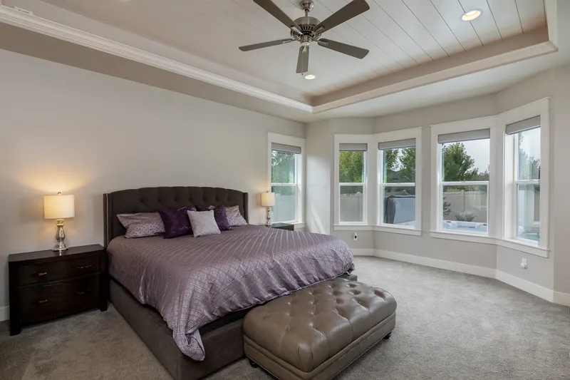 Luxurious primary bedroom in Eagle with a tufted bed, dark wood nightstands, soft gray carpeting, large bay windows, and a tray ceiling with recessed lighting.